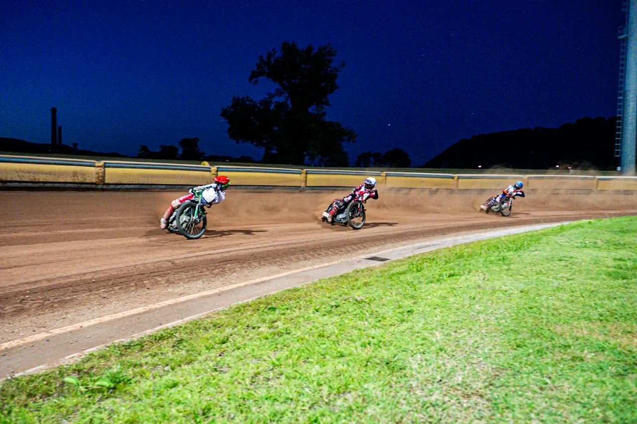 Trije dirkači speedwaya v zavoju ob mraku, nad stadionom temno modro nebo